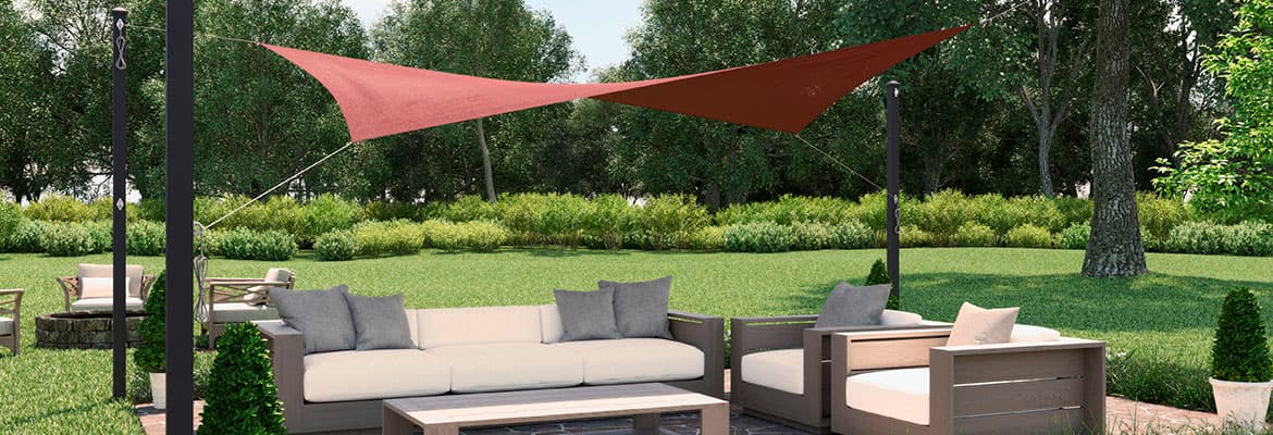 Outdoor patio scene with lush green lawn, flowers around small patio with inlaid stone pavers, wood furniture with white cushions, four large poles mounted in the ground from which hangs a Coolaroo rectangular premium shade sail kit