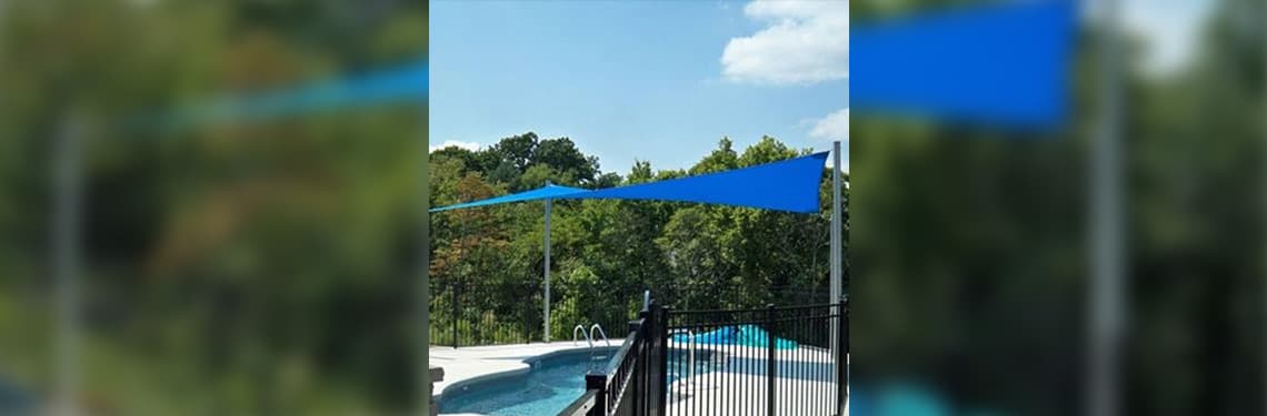 In-ground pool with white pool surround, six-foot wrought iron gate, and four tall poles mounted to the surround from which is strung a royal blue Custom Shade Sails CSS12-18REC-RB large rectangular commercial grade shade sail