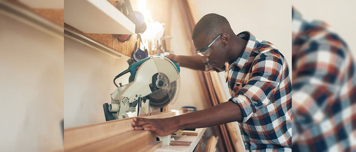 Man in plaid shirt with safety glasses, using a chop saw to cut a long piece of wood in a shop environment