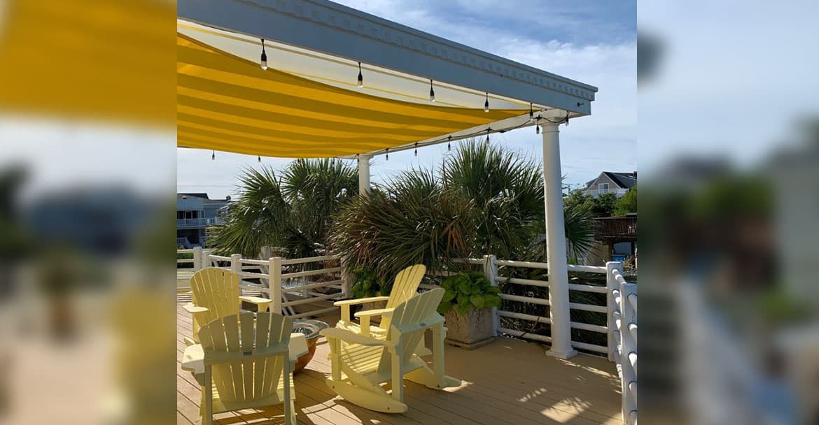 Outdoor deck scene with composite decking, white wood railing with round rails, yellow rocking Adirondack chairs around a red bowl fire pit, small white Schnauzer dog, white classic columns holding up a white wood pergola from which hangs string lights and a Custom Shade Sails CSS10-12REC-CP shade sail