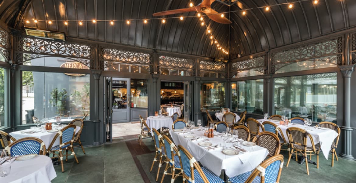 Large metal gazebo with intricate metalwork on the windows, stone tile floors, wicker chairs with white cloth covered dining tables, and string lights hanging from pergola top