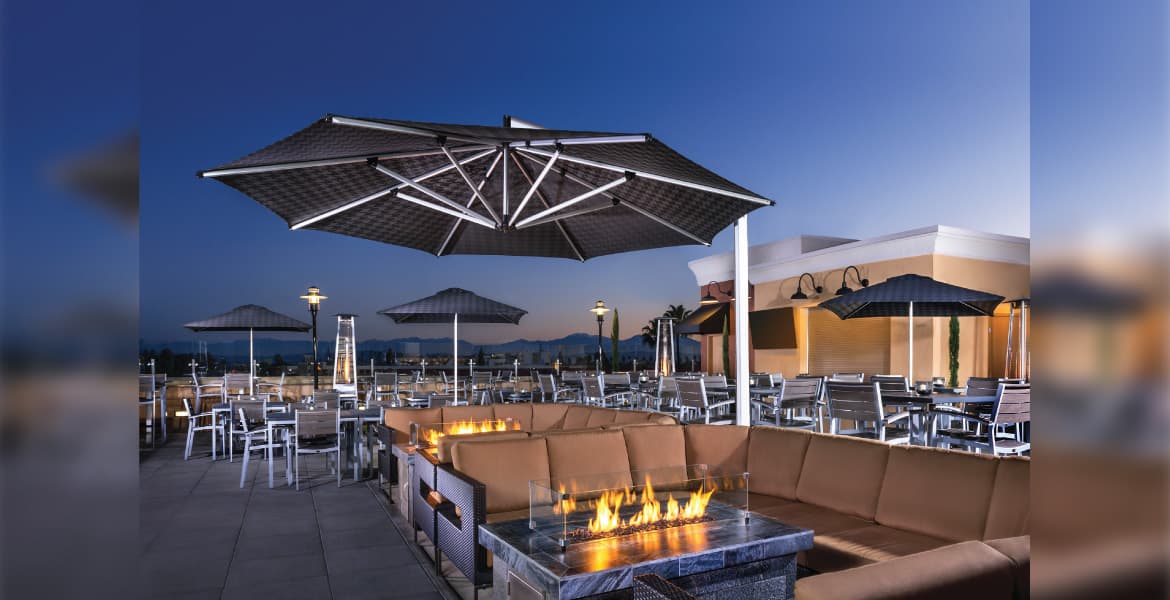 Rooftop dining scene at dusk with trendy casual pit couch seating, aluminum and wood chairs 4-top table and chairs, with glass-encased fire pit tables, portable patio heaters, multiple market umbrellas and a black Shademaker Orion OR-40 octagon sidepost umbrella