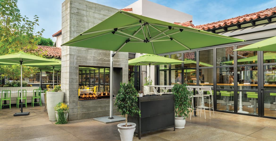 Outdoor dining area with lime-green color scheme, travertine tile floors, cinder block fireplace, modern Spanish tiled restaurant building, multiple lime-green market umbrellas and a Shademaker Polaris POLARIS-35S large square sidepost umbrella over the greeting area