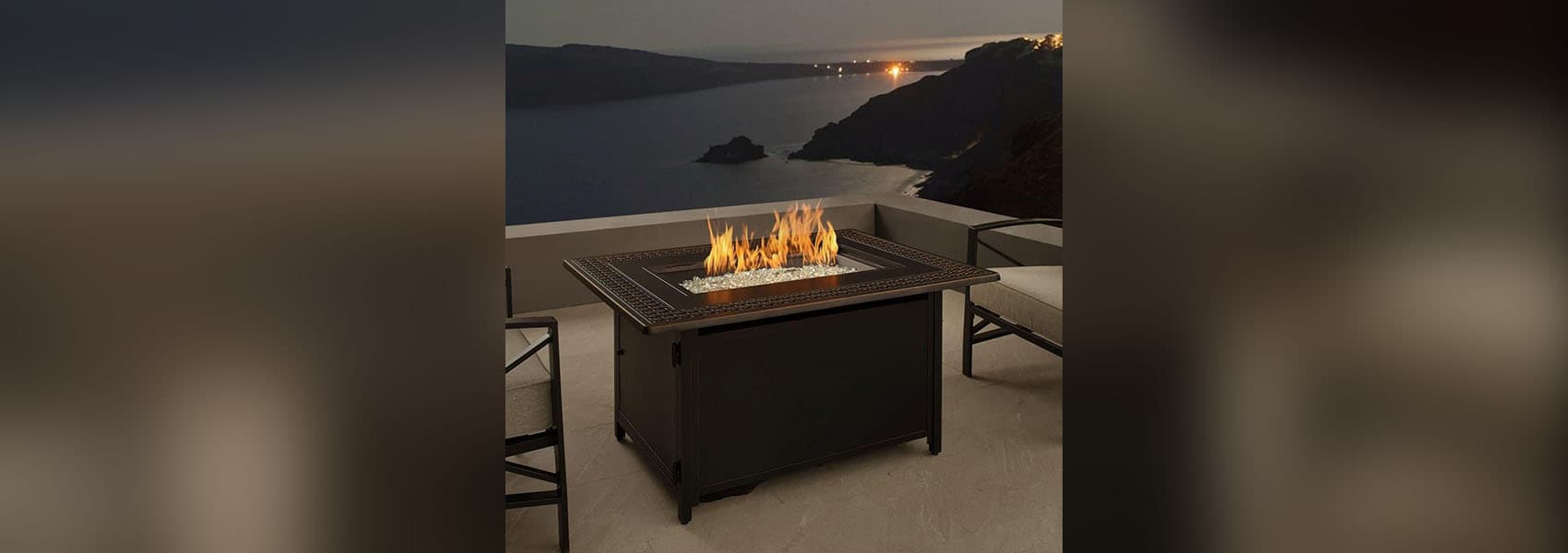 Outdoor patio scene with concrete patio with short concrete quarter wall, overlooking a small lake at sunset, with two bronze metal chairs with gray cushions on either side of a lit Fire Sense Perkins 63181 fire pit table